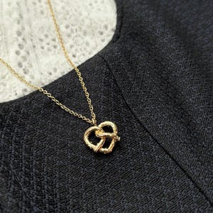 Könnte beinhalten: Eine goldfarbene Halskette mit einem kleinen, strukturierten Anhänger in Form einer Brezel. Der Anhänger hängt an einer zarten, dünnen goldfarbenen Kette. Die Metalloberfläche des Brezel-Anhängers hat ein detailliertes, leicht unebenes Finish, das das Aussehen eines gebackenen Teigknotens nachahmt.