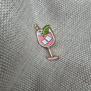 Könnte beinhalten: Ein Emaille-Anstecknadel mit einem rosa Cocktail und goldenem Umriss. Die Nadel zeigt ein Getränk mit Eiswürfeln, einer Limettenscheibe und einem gestreiften Strohhalm. Die Nadel befindet sich vor einem strukturierten, neutralen Hintergrund.