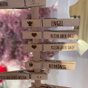 May include: A vertical wooden stand featuring multiple light-colored wooden clothespins attached to a central post. Each clothespin is laser-engraved with various German words and symbols, including "CHEF," "ENGEL," "KLEIN ABER OHO!," "BIERK&Ouml;NIG," and "PRINZESSIN."