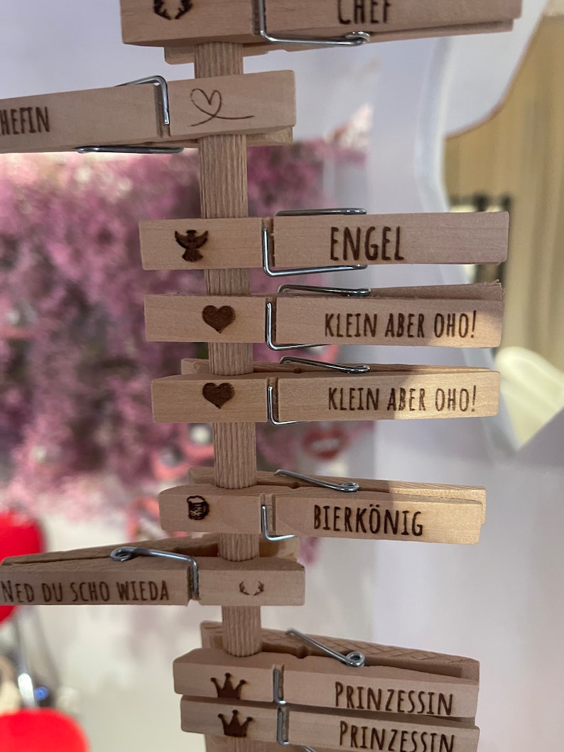 May include: A vertical wooden stand featuring multiple light-colored wooden clothespins attached to a central post. Each clothespin is laser-engraved with various German words and symbols, including "CHEF," "ENGEL," "KLEIN ABER OHO!," "BIERK&Ouml;NIG," and "PRINZESSIN."