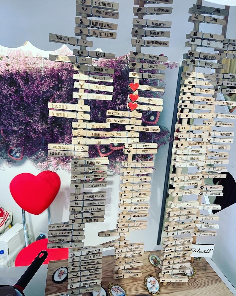 May include: Three vertical wooden displays holding numerous light-colored wooden clothespins, each engraved with German text and small icons like hearts, crowns, and pretzels. Three red heart-shaped cutouts are attached to the center display.