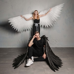 May include: A person wearing a black outfit with black angel wings stands in front of a person wearing a white outfit with white angel wings. The person with the white wings is standing on a grey background.