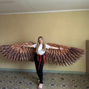 May include: Large, brown and bronze-colored angel wings are displayed on a wall behind a person. The wings are made of layered, textured material, with a gradient of colors from light brown to bronze. The person is wearing a white t-shirt, black pants, and floral shoes.