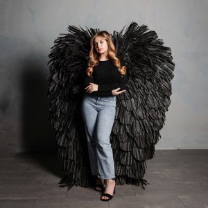 Puede incluir: Una joven con el pelo largo y rubio se encuentra delante de un gran telón de fondo de ala de ángel de plumas negras. Lleva una camisa negra de manga larga y unos vaqueros azules. Está mirando a la cámara.
