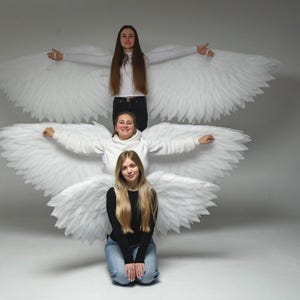 May include: Three people posing with large, white feather angel wings. The individuals are wearing casual clothing, including a white sweater, black top, and jeans. The wings are the focal point, creating a dramatic effect against a plain background.