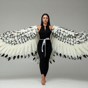 May include: Large, expansive wings featuring layered white feathers with black-tipped edges. The wings are attached to a black, sleeveless, form-fitting jumpsuit that features a front zipper and a thin white tie belt at the waist.