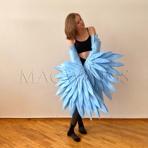 May include: A person wearing a black crop top and blue jeans is holding a pair of large, light blue wings. The wings are made of feathers and are spread out behind the person.