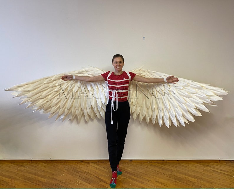 May include: A person wearing a red and white striped shirt and black trousers is holding out large white angel wings. The wings are made of feathers and are very detailed.