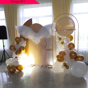 May include: A gold and white balloon arch with a white feather angel wing backdrop. The arch is decorated with gold and white balloons and a sign that says "You are my sunshine".