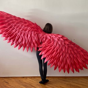 May include: A pair of large, bright pink angel wings made of feathers. The wings are being worn by a person standing in front of a white wall.