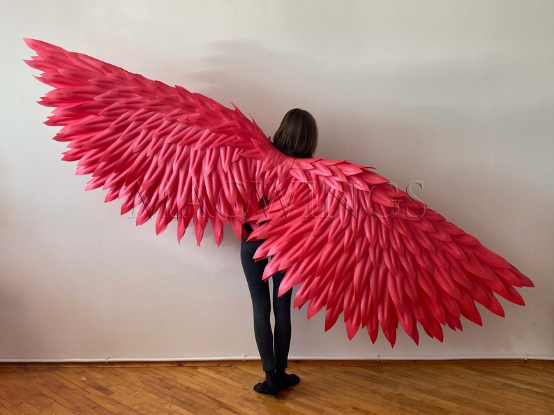 May include: A pair of large, bright pink angel wings made of feathers. The wings are being worn by a person standing in front of a white wall.