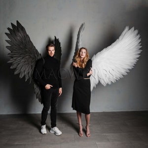 May include: Two people wearing angel wings, one black and one white. The person with the black wings is wearing a black turtleneck and black trousers. The person with the white wings is wearing a black dress. Both people are standing in front of a grey wall.