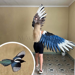May include: A person wearing a costume with large blue and white wings, resembling a magpie. The wings are made of feathers and are attached to the person's back. The person is standing on a tiled floor. A small magpie is shown in the lower left corner of the image.