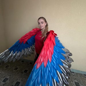 以下が含まれることがあります： 大きなオウムのカラフルな翼をつけた人。翼は赤、青、銀の色合いで、羽の層状のデザインが特徴です。その人は赤いシャツを着ています。翼は大きく広げられ、鮮やかな色と細部が示されています。