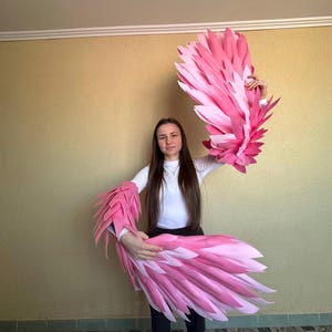 May include: A pair of pink and light pink flamingo wings. The wings are made of layered, textured material, with a feathery appearance. The wings are held up, showcasing their size and design. The overall theme is costume or cosplay.