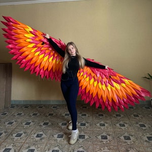 May include: A person wearing black clothing poses with large, bright red and orange wings made of paper. The wings are shaped like a bird's wings and have a layered, feathery design.