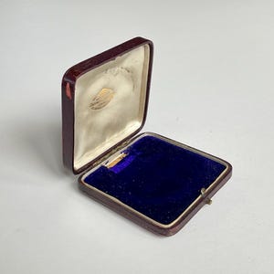 May include: An open, rectangular jewellery box with a burgundy exterior and a blue velvet interior. The inside lid is lined with off-white satin. A small, gold-coloured emblem is visible on the satin lining. The box is open, revealing its interior.