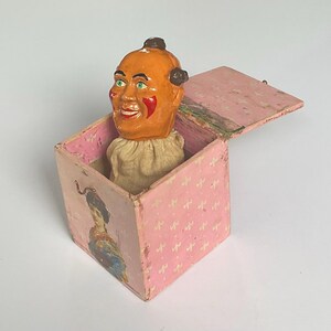 May include: A vintage Jack-in-the-box toy with a pink box and a smiling clown head. The box has a pink background with a repeating pattern of gold crosses. The clown has orange skin, red cheeks, and a white ruffled collar.