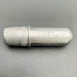 May include: A silver metal thimble with the words "SOAP" and "HUDSON'S" engraved on the side. The thimble is attached to a small metal tube with the words "USE" and "SOAP" engraved on it.