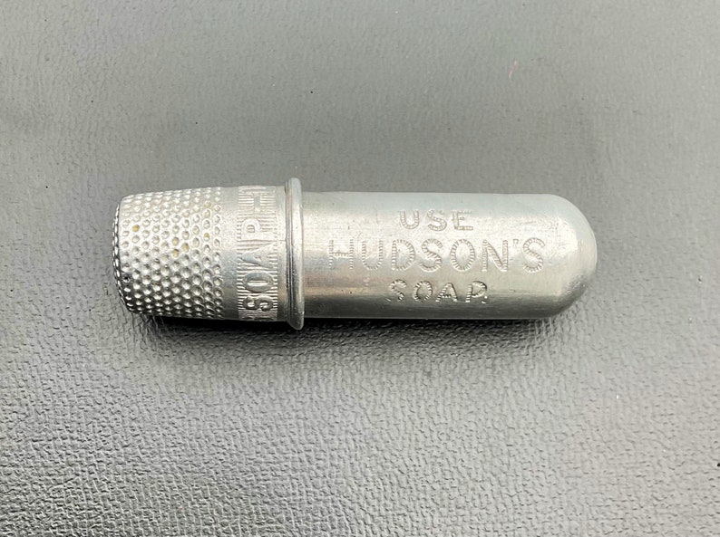 May include: A silver metal thimble with the words "SOAP" and "HUDSON'S" engraved on the side. The thimble is attached to a small metal tube with the words "USE" and "SOAP" engraved on it.