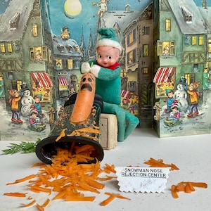 Peut inclure: Une scène de Noël fantaisiste avec une poupée elfe verte râpant un nez de carotte pour un bonhomme de neige. La carotte est placée sur une râpe, avec des morceaux de carottes râpées éparpillés. Le texte "SNOWMAN NOSE REJECTION CENTER" est visible. Un décor de village d'hiver complète la scène.