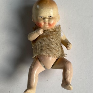 May include: A vintage doll with a painted face, rosy cheeks, and a burlap sackcloth vest. The doll's limbs are in a bent position, and it has a light skin tone. The doll's hair is painted brown, and it has a serious expression.