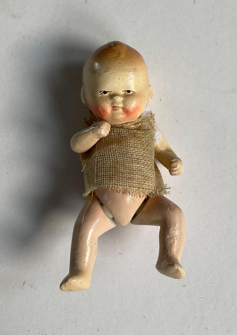 May include: A vintage doll with a painted face, rosy cheeks, and a burlap sackcloth vest. The doll's limbs are in a bent position, and it has a light skin tone. The doll's hair is painted brown, and it has a serious expression.