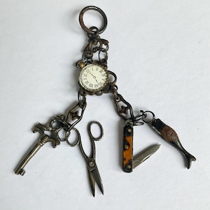 Puede incluir: Un soporte para dijes tipo châtelaine vintage con una esfera de reloj circular central con números romanos, suspendido de un anillo de metal. Incluye cuatro dijes colgantes: una llave de metal ornamentada, unas tijeras pequeñas, una navaja y un pez.