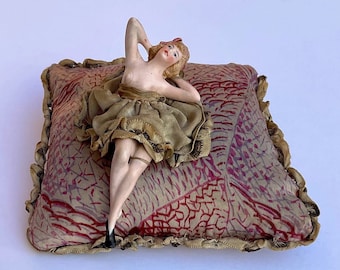 May include: A porcelain figurine of a woman in a gold dress lying on a red and gold patterned pillow. The figurine is nude from the waist up.