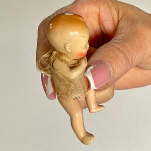 May include: A vintage bisque doll with a peach-colored head and rosy cheeks. The doll is wearing a tan fabric garment. The doll's legs are extended, and it is held in a hand.