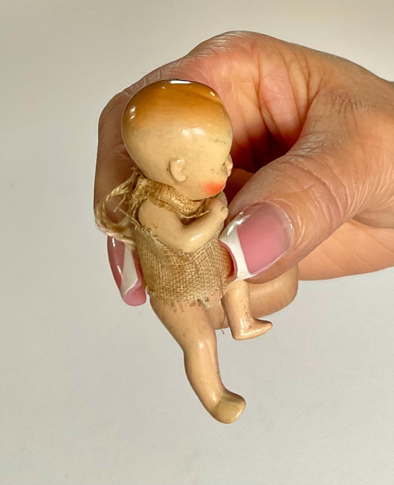 May include: A vintage bisque doll with a peach-colored head and rosy cheeks. The doll is wearing a tan fabric garment. The doll's legs are extended, and it is held in a hand.