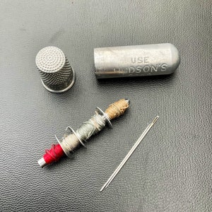 May include: A silver thimble, a small spool of thread with red, green, and brown thread, a silver needle, and a silver case with the text "USE HUDSON'S" on it.
