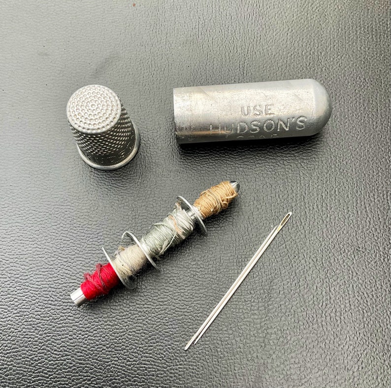 May include: A silver thimble, a small spool of thread with red, green, and brown thread, a silver needle, and a silver case with the text "USE HUDSON'S" on it.