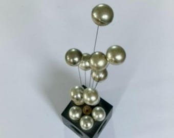 Könnte beinhalten: Eine dekorative Skulptur mit mehreren metallischen Kugeln unterschiedlicher Größe. Die Kugeln sind an dünnen Drähten befestigt, die von einer dunklen quadratischen Basis aufsteigen. Die Kugeln haben eine reflektierende, perlmuttartige Oberfläche.