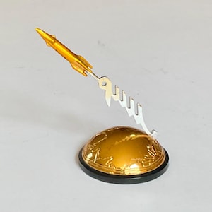 Puede incluir: Una figura de cohete dorada con un diseño de texto estilizado plateado, montada sobre una base de globo dorada con un borde negro. El cohete está inclinado hacia arriba, sugiriendo vuelo.