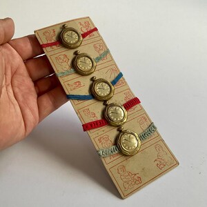 以下が含まれることがあります： 赤、青、グレーのリボンで取り付けられた、5つの小さな金色の懐中時計が特徴のヴィンテージカード。カードには動物や人々の赤いイラストが描かれています。時計は手で持たれています。