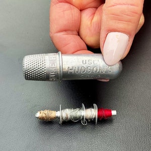 May include: A silver thimble with the words "Use Hudson's Soap" engraved on the side. A small spool of thread with red, brown, and gray thread is also pictured.