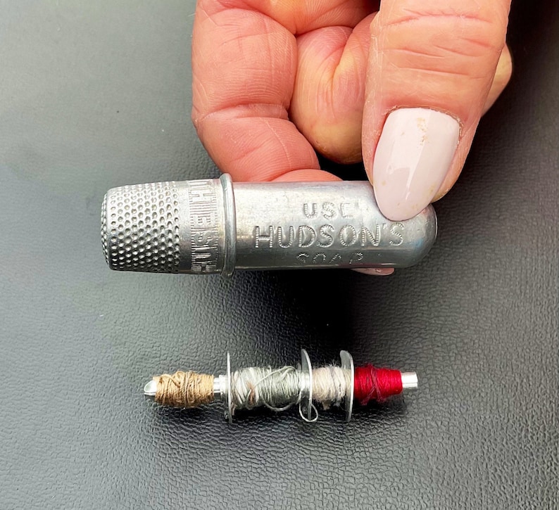 May include: A silver thimble with the words "Use Hudson's Soap" engraved on the side. A small spool of thread with red, brown, and gray thread is also pictured.