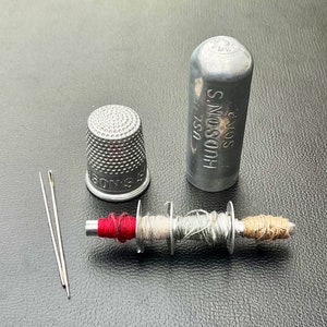 May include: A silver thimble, a small spool of thread with red, white, and brown thread, two needles, and a silver container with the text "HUDSON'S SOAP" on it.