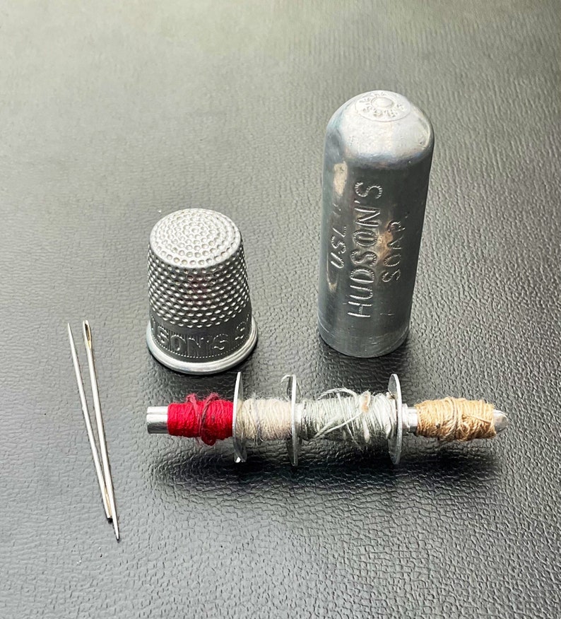 May include: A silver thimble, a small spool of thread with red, white, and brown thread, two needles, and a silver container with the text "HUDSON'S SOAP" on it.
