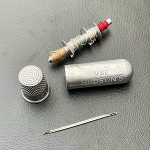 May include: A silver metal thimble with a textured surface. A silver metal spool with a red, white, and brown thread. A silver metal tube with the text "USE HUDSON'S" and a silver metal needle.