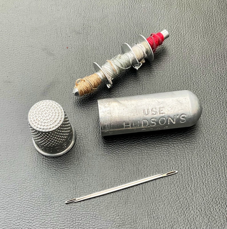 May include: A silver metal thimble with a textured surface. A silver metal spool with a red, white, and brown thread. A silver metal tube with the text "USE HUDSON'S" and a silver metal needle.