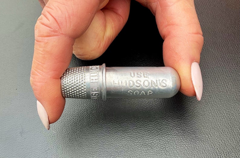 May include: A silver thimble attached to a small metal container with the text "USE HUDSON'S SOAP" engraved on it.