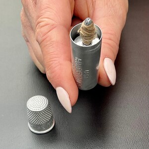 May include: A silver thimble with a textured surface. A silver metal container with the text "HUDSON'S" and "USA" is open and contains a small spool of thread.