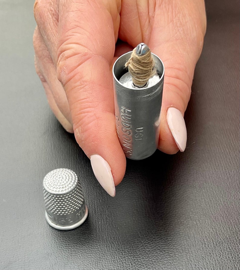 May include: A silver thimble with a textured surface. A silver metal container with the text "HUDSON'S" and "USA" is open and contains a small spool of thread.