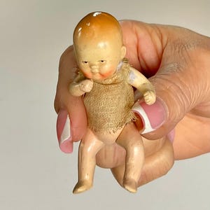 May include: A small, vintage doll with a painted face and limbs. The doll has a tan-colored head with a darker top, rosy cheeks, and is wearing a burlap sack. The doll is held in a hand with a pink and white manicure.