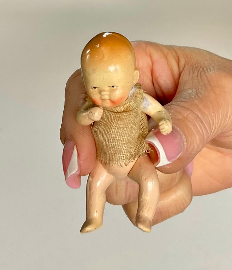 May include: A small, vintage doll with a painted face and limbs. The doll has a tan-colored head with a darker top, rosy cheeks, and is wearing a burlap sack. The doll is held in a hand with a pink and white manicure.