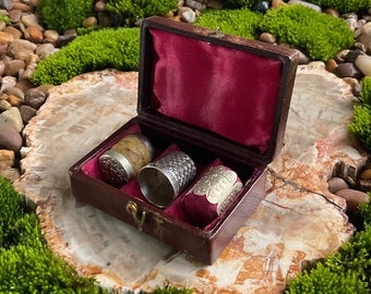 May include: A vintage wooden box with a red velvet interior containing three thimbles. Two of the thimbles are silver with intricate designs, and one is made of wood with a brown and white pattern.