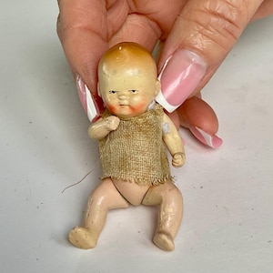 May include: A small, vintage doll with a painted face and limbs. The doll has a tan burlap top and is posed in a seated position. The doll's hair is painted a light brown color. The doll is being held by a person.