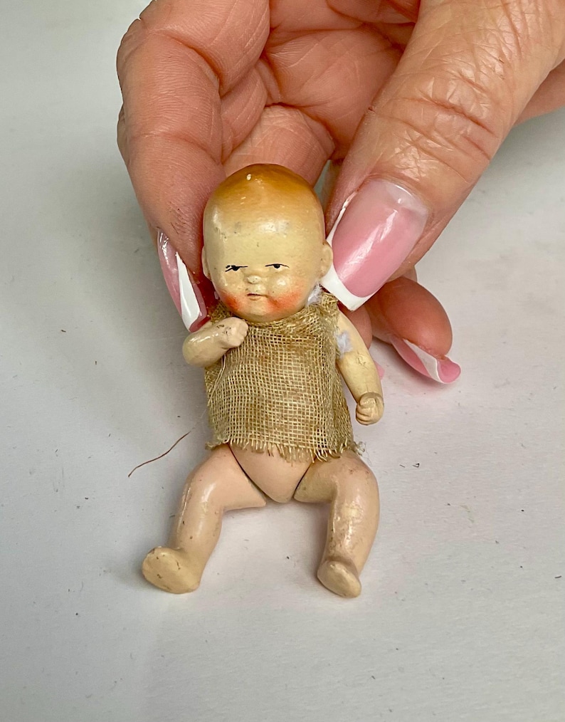 May include: A small, vintage doll with a painted face and limbs. The doll has a tan burlap top and is posed in a seated position. The doll's hair is painted a light brown color. The doll is being held by a person.